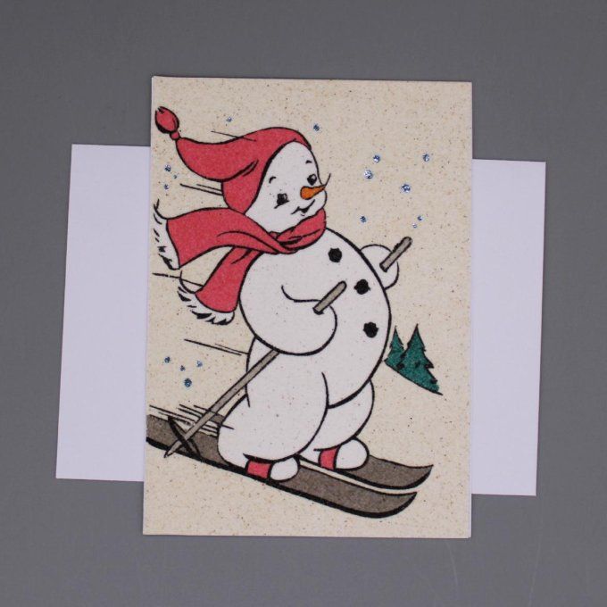 Carte de vœux représentant un bonhomme de neige qui skie, réalisée en sable coloré.