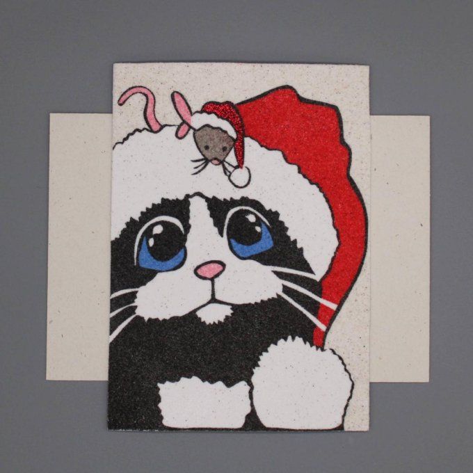 Carte de Noël en sable représentant un chat noir et blanc avec un bonnet de Noël, surmonté d’une pet