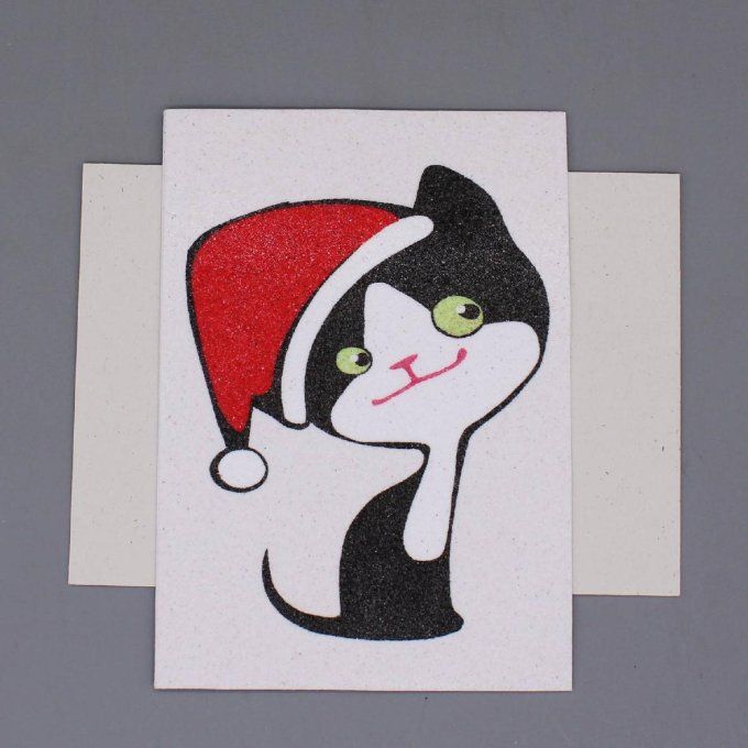 Carte de Noël en sable montrant un chat noir et blanc avec un bonnet rouge scintillant.