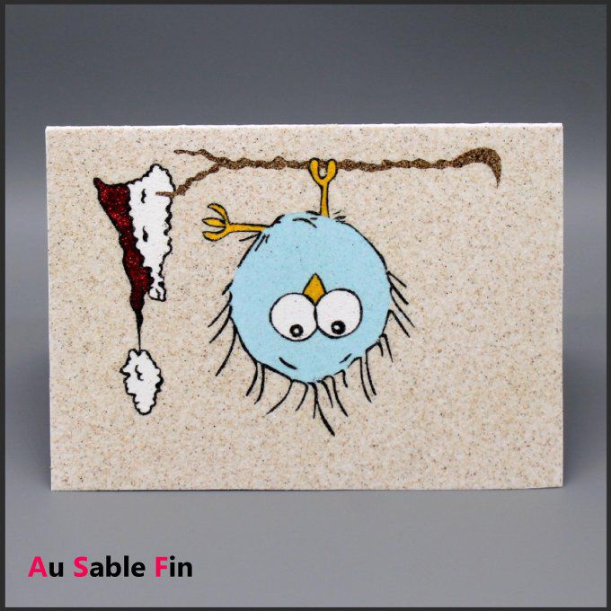 Carte de Noël en sable montrant un petit oiseau bleu suspendu à une branche, son bonnet rouge tomban