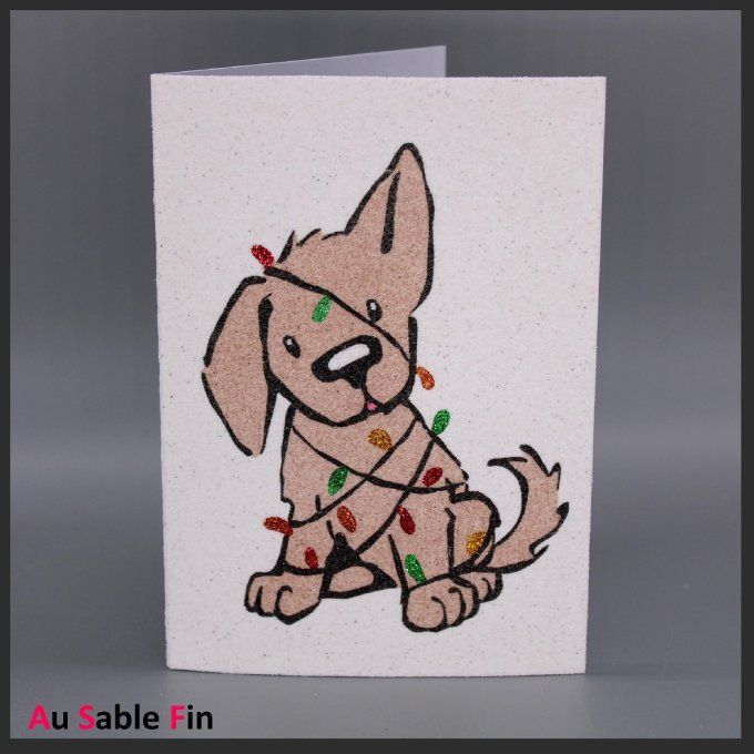 Carte de Noël en sable montrant un chiot beige enveloppé d’une guirlande lumineuse.